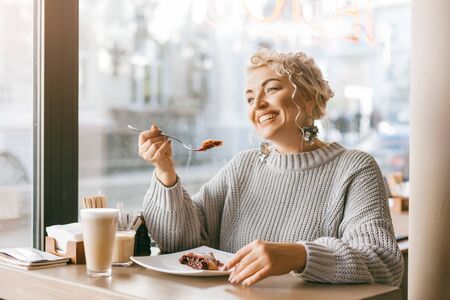 Positive beautiful plus size woman drinking coffee and eating sweet dessert by the window in city street cafeの写真素材