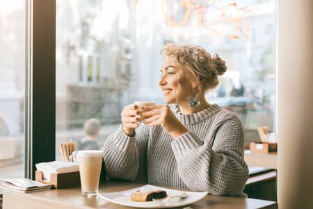 Positive beautiful plus size woman drinking coffee and eating sweet dessert by the window in city street cafeの写真素材