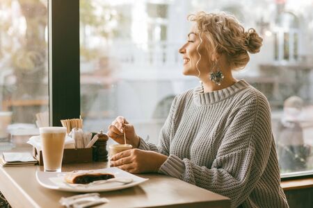 Positive beautiful plus size woman drinking coffee and eating sweet dessert by the window in city street cafeの写真素材