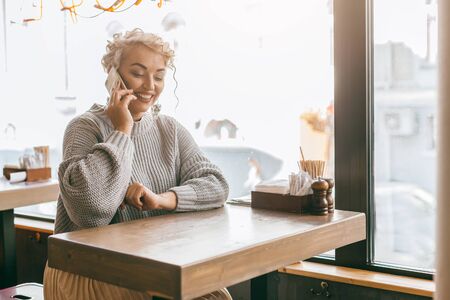 Positive beautiful plus size woman using smart phone while sitting by the window in city street cafeの写真素材