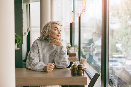 Positive beautiful plus size woman waiting for meal while sitting by the window in city street cafeの写真素材