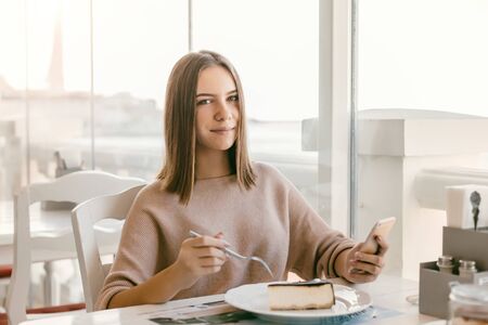 Cute teen girl using smart phone and enjoying sweet dessert while spending time in bright cafeの写真素材