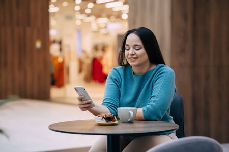 Beautiful plus size woman using mobile phone and eating dessert with coffe while resting in cafe inside city mallの写真素材