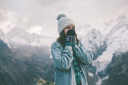 Young traveler girl in gloves and hat standing over snowy mountain peaks and warming cold hands. Winter travelling scene, wanderlust concept.の写真素材