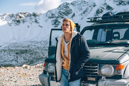 Young guy standing alone near 4x4 vehicle and enjoying view of mountains. Winter offroad trip on rental car to wild nature.の写真素材