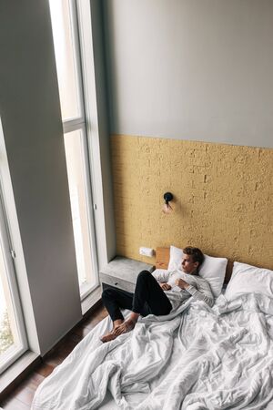 Young man relaxing on comfy bed and looking at window in modern hotel roomの写真素材