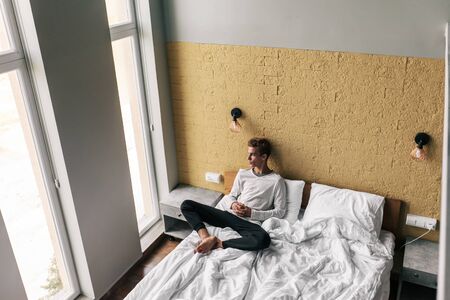 Young man relaxing on comfy bed and looking at window in modern hotel roomの写真素材