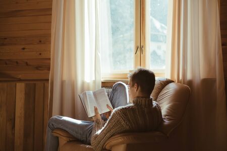Young man in warm sweater reading book while relaxing on armchair by the window inside cozy log cabinの写真素材