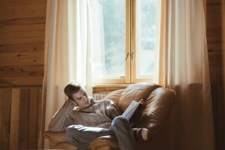 Young man in warm sweater reading book while relaxing on armchair by the window inside cozy log cabinの写真素材