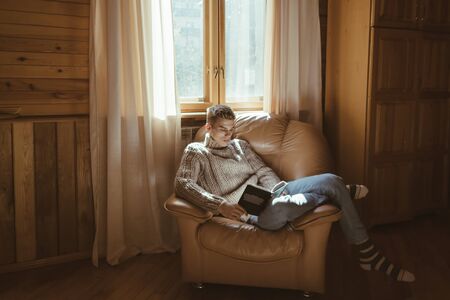 Young man in warm sweater reading book while relaxing on armchair by the window inside cozy log cabinの写真素材