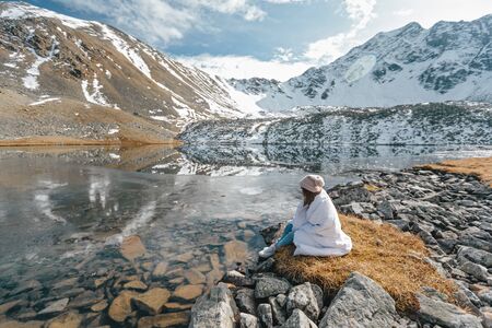 Young girl covered in blanket relaxing in wild nature with a view on beautiful landscape in cold season. Dreamy wanderlust concept scene. Spending perfect winter vacations by lake in mountains.の写真素材
