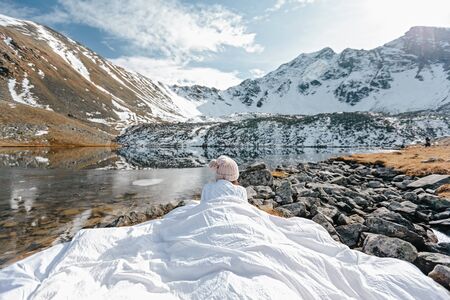 Young girl relaxing in comfy bed in wild nature with a view on beautiful landscape in cold season. Dreamy wanderlust concept scene. Spending perfect winter vacations by lake in mountains.の写真素材