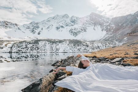 Young girl relaxing in comfy bed in wild nature with a view on beautiful landscape in cold season. Dreamy wanderlust concept scene. Spending perfect winter vacations by lake in mountains.の写真素材