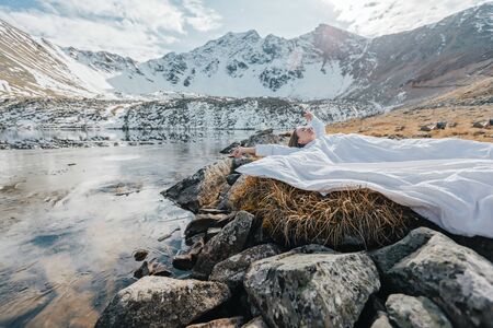 Young girl relaxing in comfy bed in wild nature with a view on beautiful landscape in cold season. Dreamy wanderlust concept scene. Spending perfect winter vacations by lake in mountains.の写真素材