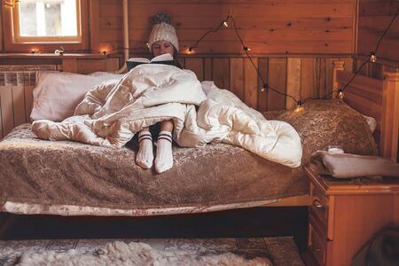 Young girl relaxing and reading book under warm duvet on cozy bed in log cabin in winterの写真素材