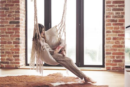 Beautiful young woman, which is wearing cashmere nightwear clothes, reading novel on hammock in...