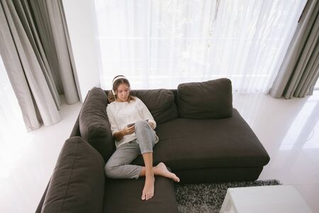 Teen girl using tablet pc while relaxing on couch in hotel room in front of big window with curtainsの写真素材
