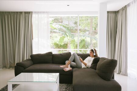 Teen girl using tablet pc while relaxing on couch in hotel room in front of big window with curtainsの写真素材