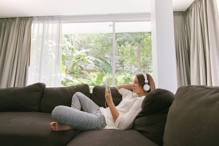 Teen girl using tablet pc while relaxing on couch in hotel room in front of big window with curtainsの写真素材