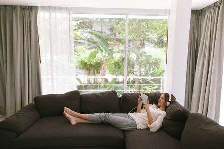Teen girl using tablet pc while relaxing on couch in hotel room in front of big window with curtainsの写真素材