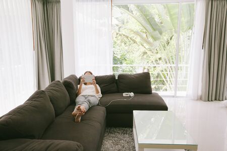 Teen girl using tablet pc while relaxing on couch in hotel room in front of big window with curtainsの写真素材