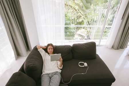 Teen girl using tablet pc while relaxing on couch in hotel room in front of big windowの写真素材