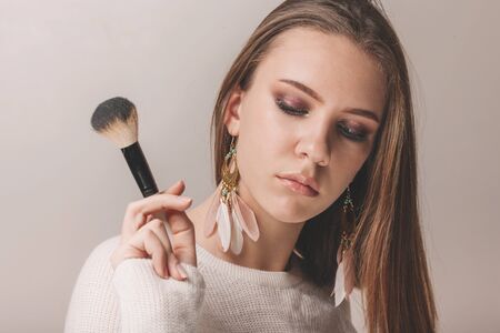 Studio portrait of beautiful teen model with stylish makeup holding powder or contour brush on light...