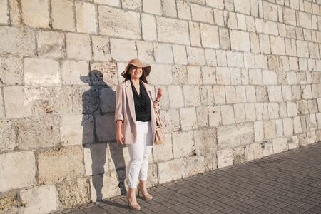 Young stylish woman wearing neutral blazer and hat walking on the city street in evening. Casual fashion, elegant look. Plus size model.の写真素材