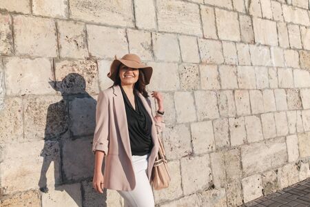 Young stylish woman wearing neutral blazer, hat and handbag walking on the city street in spring. Casual fashion, elegant look. Plus size model.の写真素材