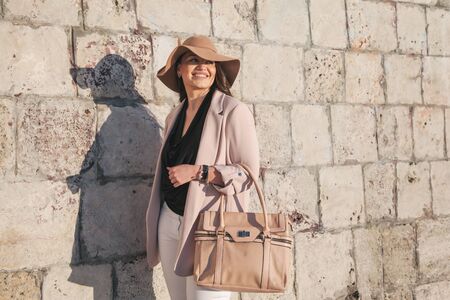 Young stylish woman wearing neutral blazer, hat and handbag walking on the city street in spring. Casual fashion, elegant look. Plus size model.の写真素材