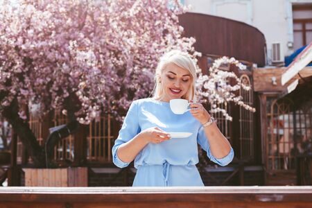 Beauitful plus size model wearing blue dress and trendy sunglasses drinking morning coffee under pink blooming spring tree outdoor in city cafeの写真素材
