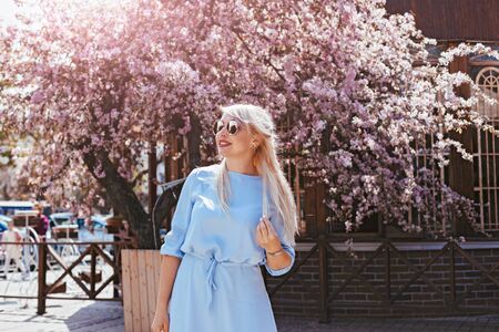 Beauitful plus size model wearing blue dress and trendy sunglasses enjoying smell of pink blooming spring tree outdoor on the city street.の写真素材