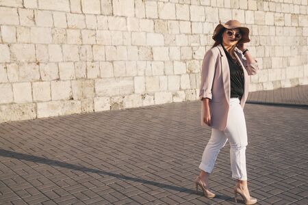 Young stylish woman wearing neutral blazer and hat walking on the city street in evening. Casual fashion, elegant look. Plus size model.の写真素材