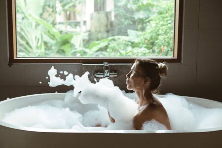 Woman relaxing with pleasue in foam bath with bubbles in modern hotel dark bathroom by big window with tropical viewの写真素材
