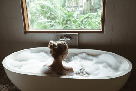Back view of woman relaxing with pleasue in foam bath with bubbles in modern hotel dark bathroom by...