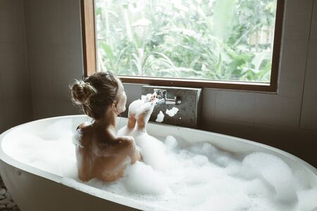 Back view of woman relaxing with pleasue in foam bath with bubbles in modern hotel dark bathroom by big windowの写真素材