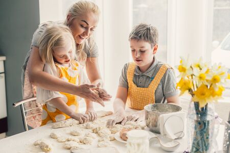 Beautiful blond mom teaching her two children cooking on the kitchen. Parent making everyday breakfast together with kids. Family at home lifestyle photo.の写真素材