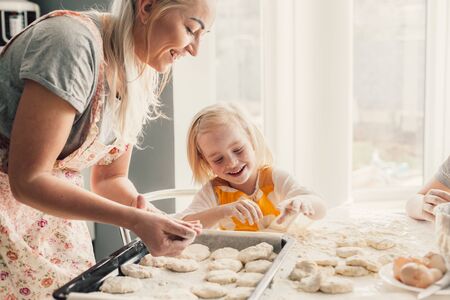 Beautiful blond mom teaching her daughter cooking on the kitchen. Parent making everyday breakfast together with child. Family at home lifestyle photo.の写真素材