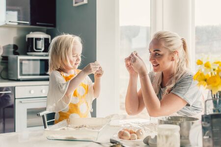 Beautiful blond mom teaching her daughter cooking on the kitchen. Parent making everyday breakfast together with child. Family at home lifestyle photo.の写真素材