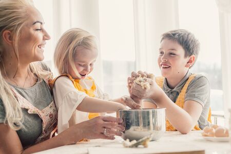 Beautiful blond mom teaching her two children cooking on the kitchen. Parent making everyday breakfast together with kids. Family at home lifestyle photo.の写真素材