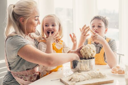 Beautiful blond mom teaching her two children cooking on the kitchen. Parent making everyday breakfast together with kids. Family at home lifestyle photo.の写真素材