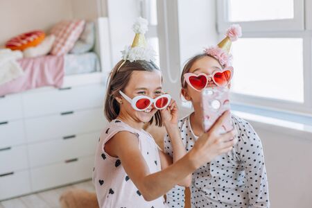 Two children playing together, taking selfie on smartphone, having fun in bright girly playroom. Kids paljamas party in white bedroom interior.の写真素材