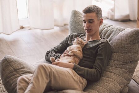 Owner playing with cat while relaxing on modern couch in living room interior. Young man resting with pet in soft chair at home.の写真素材