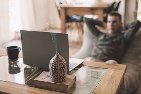 Handsome teenage guy working on laptop while relaxing on modern couch in living room interior. Young man resting in soft chair at home using aroma incence stick.の写真素材