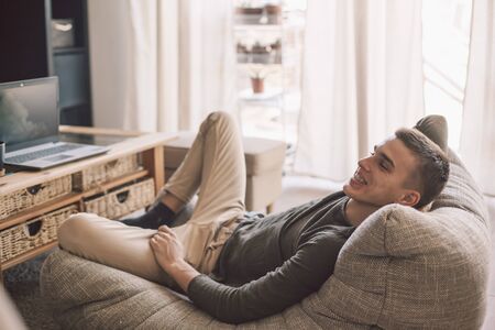Handsome teenage guy with dental braces smiling while relaxing on modern couch in living room interior. Happy man resting in soft chair at home.の写真素材