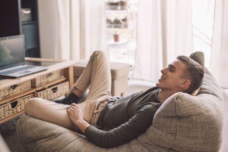 Handsome teenage guy with dental braces smiling while relaxing on modern couch in living room interior. Happy man resting in soft chair at home.の写真素材