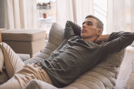 Handsome teenage guy relaxing on modern couch in living room interior. Young man resting in soft chair at home.の写真素材