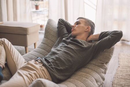 Handsome teenage guy relaxing on modern couch in living room interior. Young man resting in soft chair at home.の写真素材