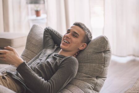 Handsome teenage guy with dental braces smiling while relaxing on modern couch in living room...