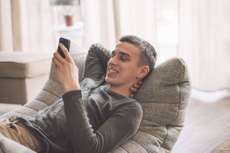 Handsome teenage guy with dental braces smiling and using smartphone while relaxing on modern couch in living room interior. Happy man resting in soft chair at home.の写真素材